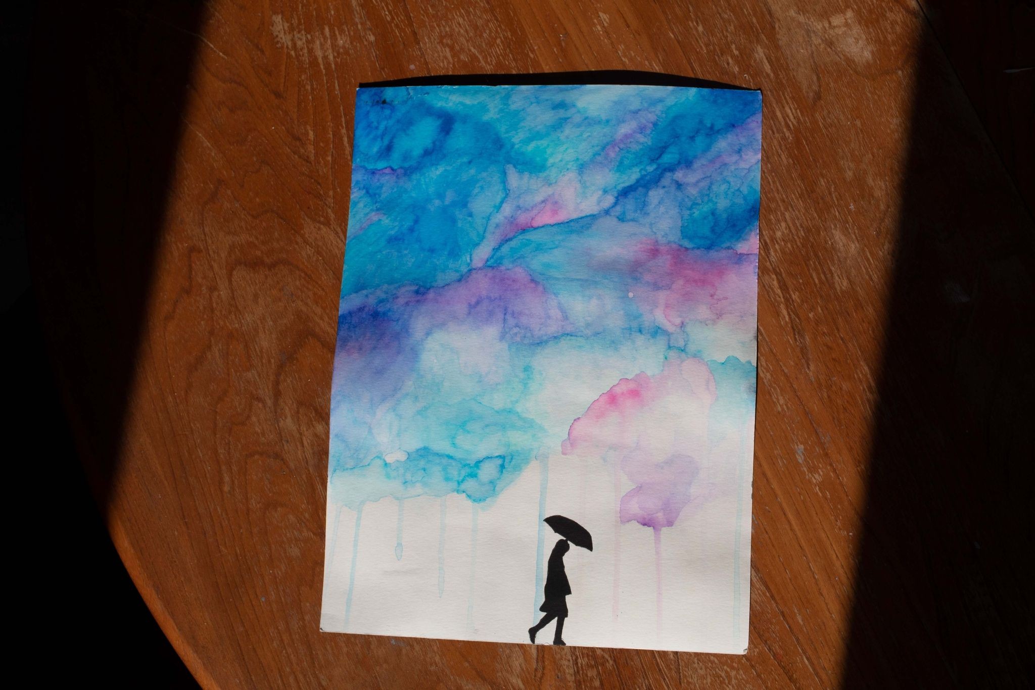 Product image of a watercolour painting of blue and pink clouds raining onto the silhouette of a person holding an umbrella, viewed from farther away