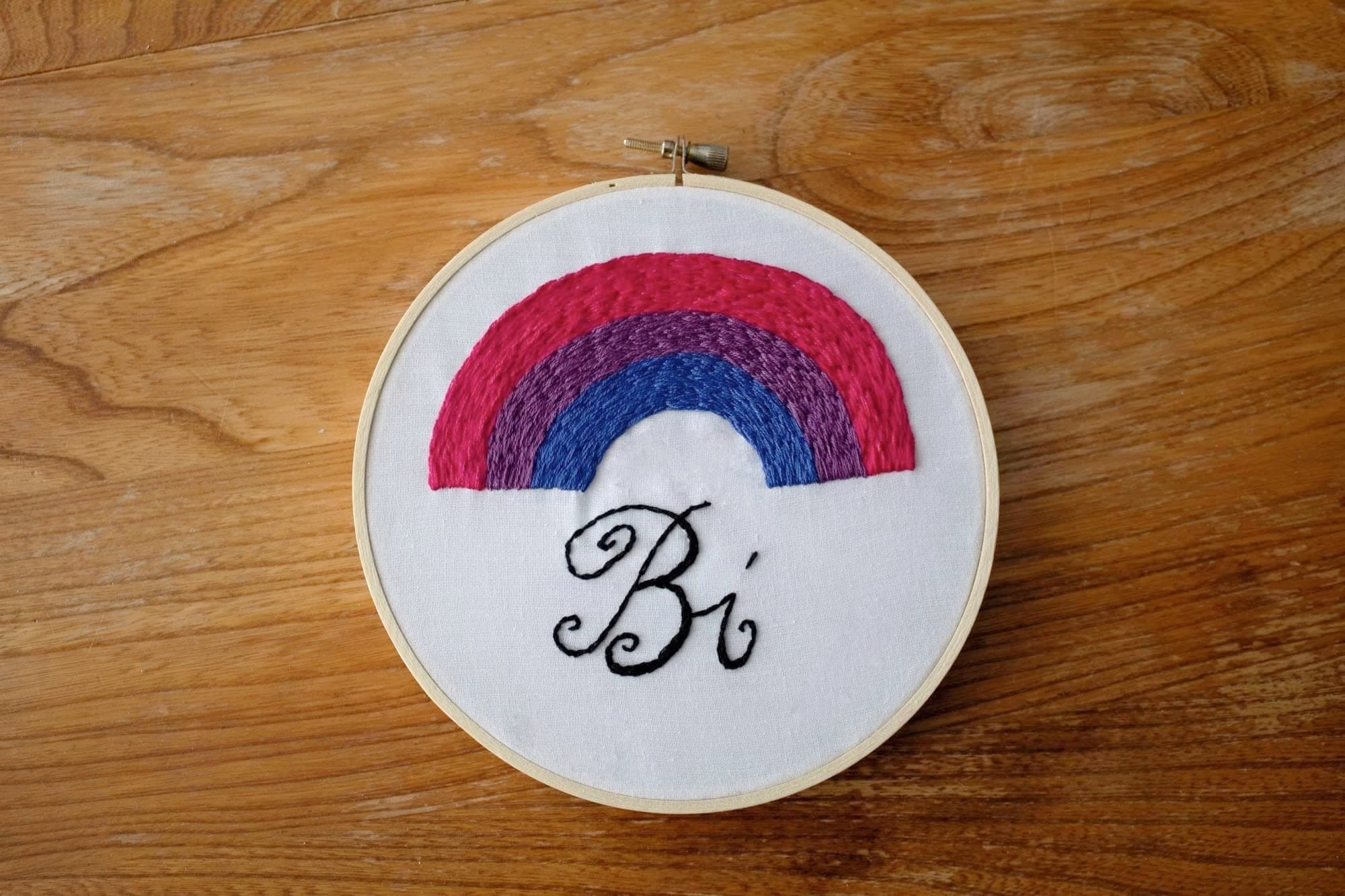 Product image of embroidered rainbow in the bi flag colours with "Bi" written underneath in an italic font on a white fabric background, the whole thing set against a wooden backdrop