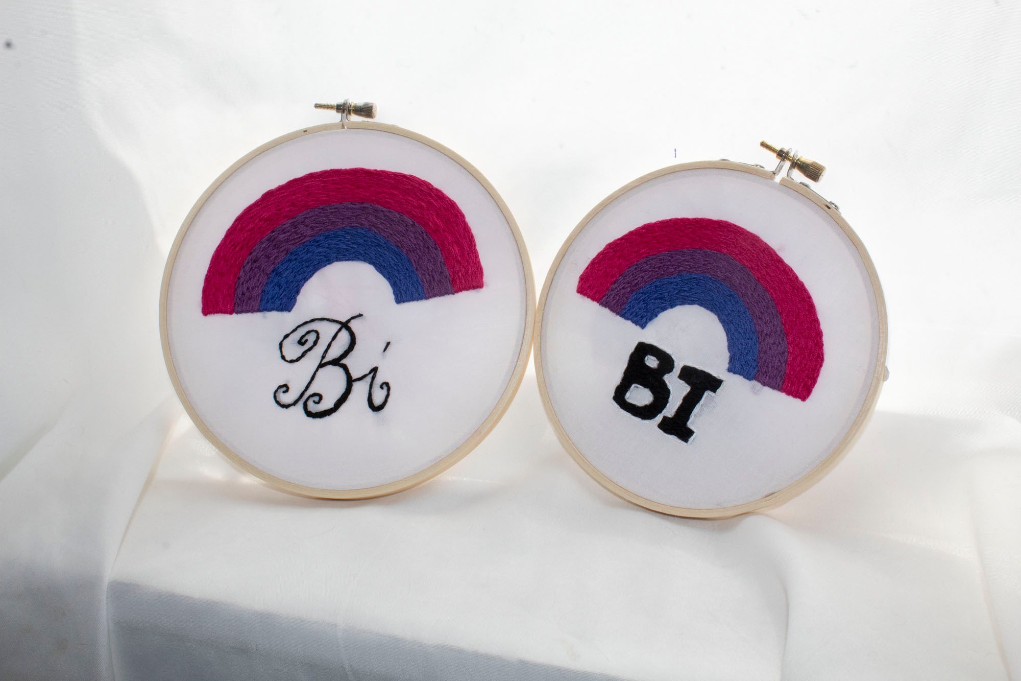 Product image of both styles of Bi Pride embroideries set against a white curtain