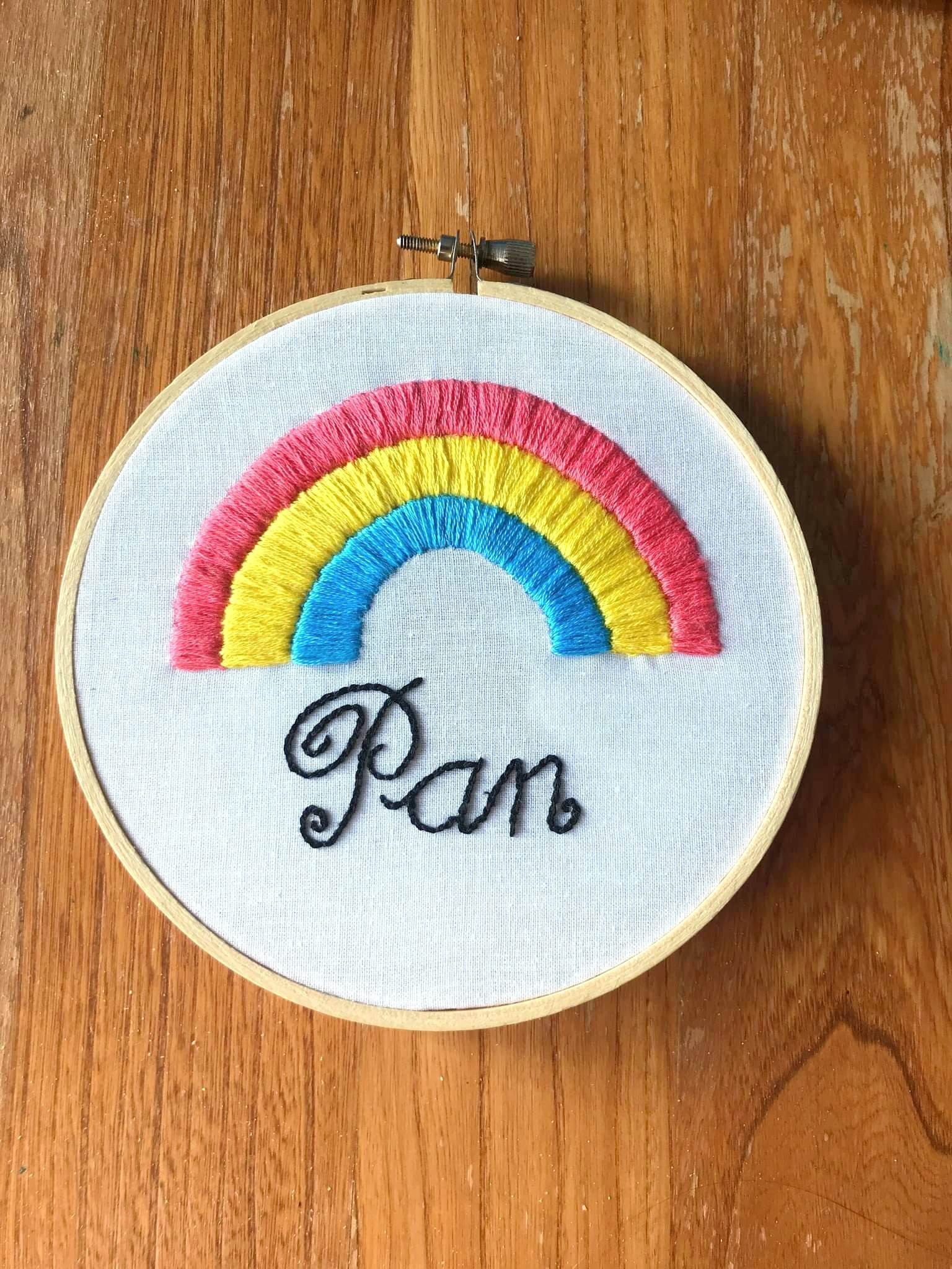 Product image of embroidered rainbow in the pansexual flag colours, with the word "pan" written in italics at the bottom in black, the whole embroidered on white fabric and set against a wooden backdrop