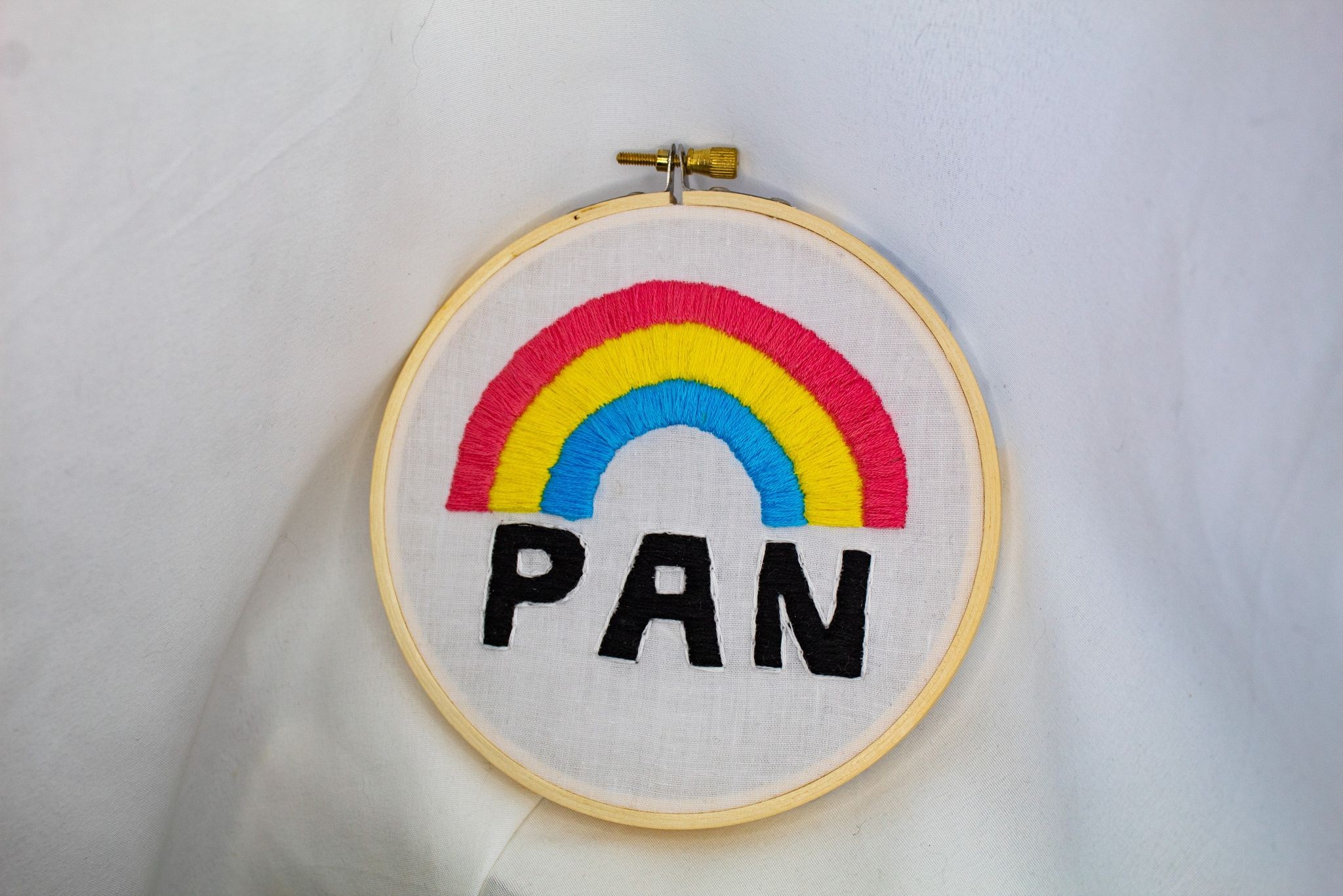 Product image of a rainbow in the pan flag colours with the word "pan" written in bold black letters underneath, embroidered on white fabric, the whole set against a white curtain