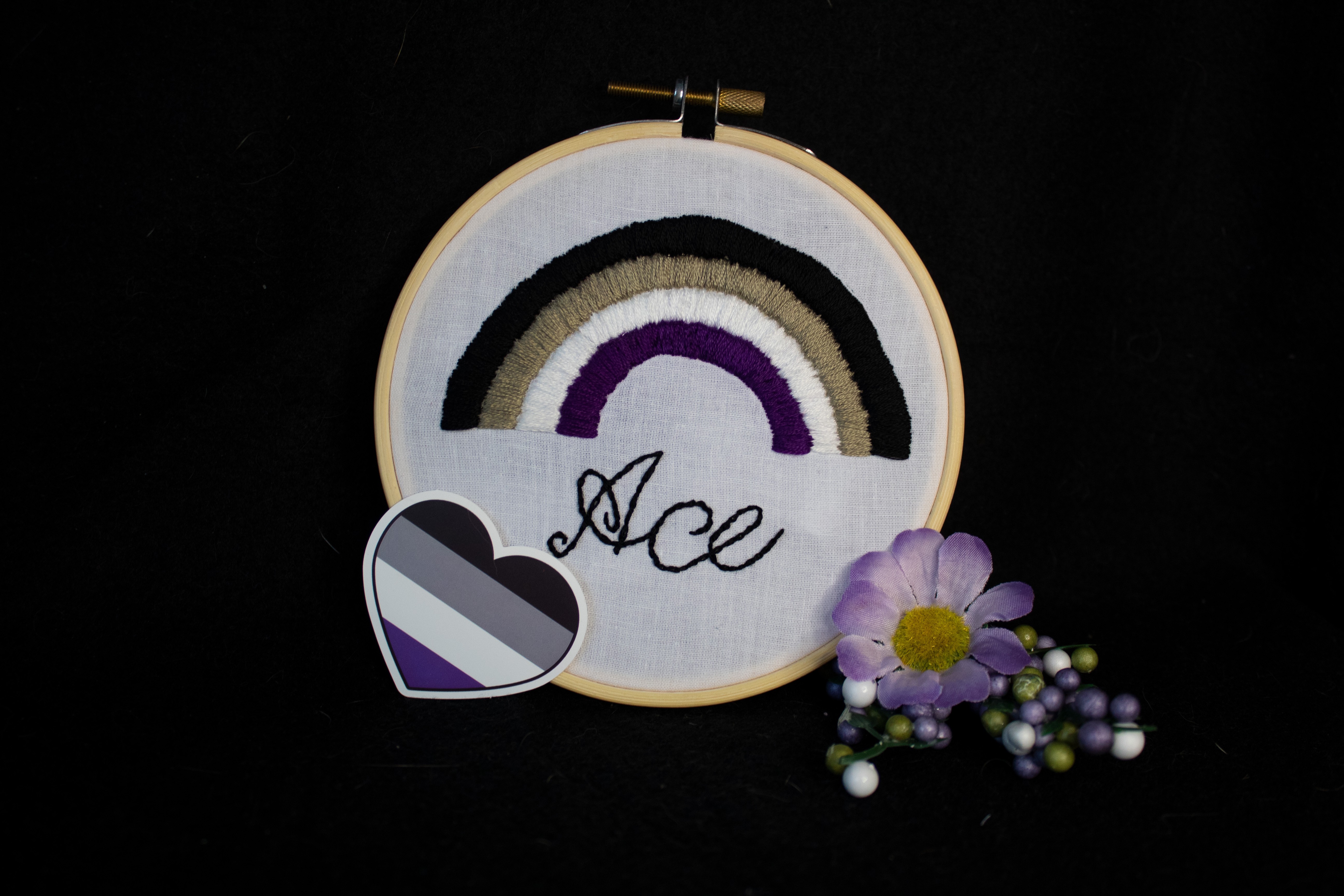 Product image of the ace embroidery set against a black backdrop, with a sprig of purple and green buds and one purple  one one side, and a heart-shaped sticker of the asexual flag on the other side