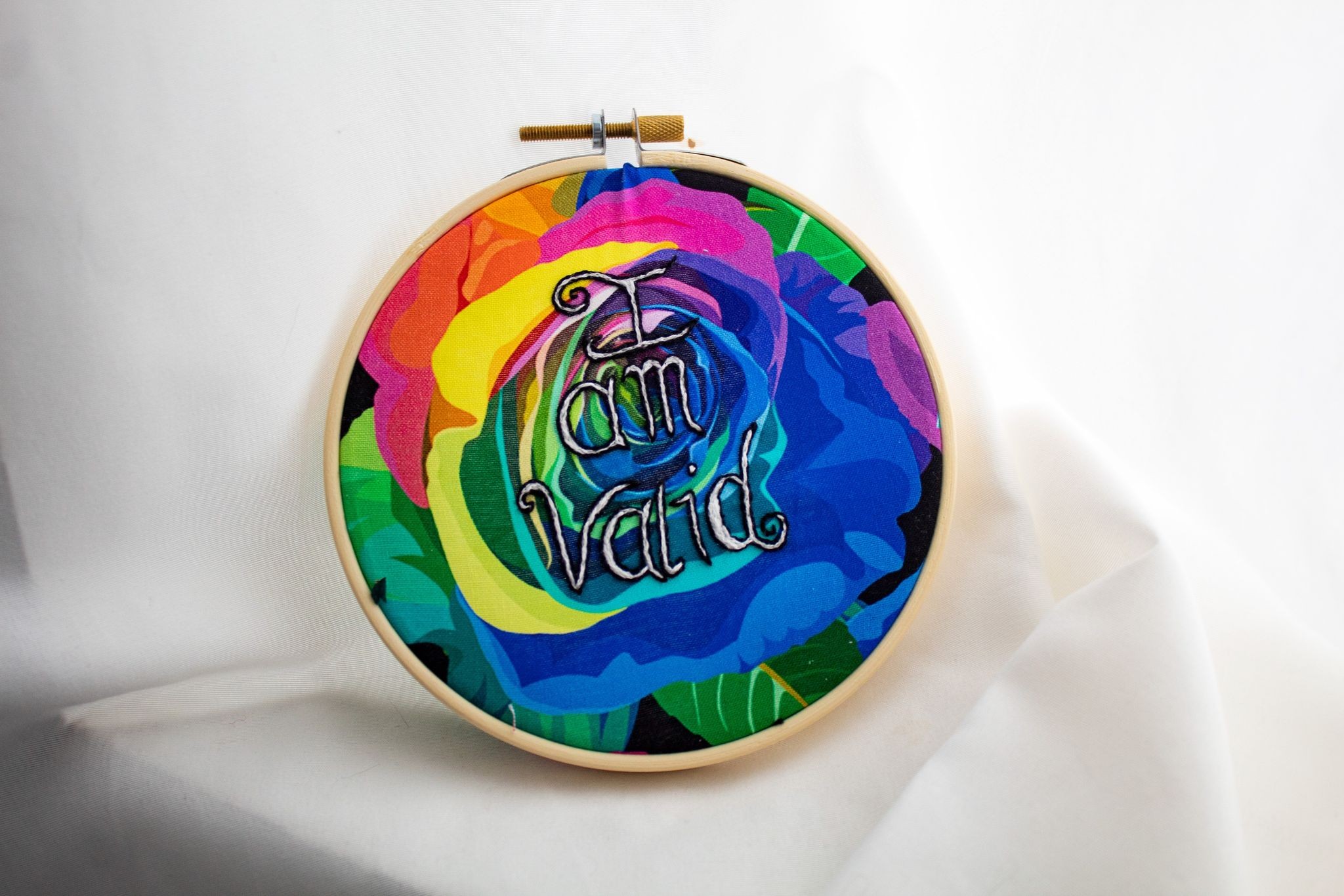 Product image of an embroidery featuring the words "I am valid"  in white thread with black outline embroidered over  rainbow-coloured rose-printed fabric, the whole in a wooden hoop and set against a white fabric backdrop