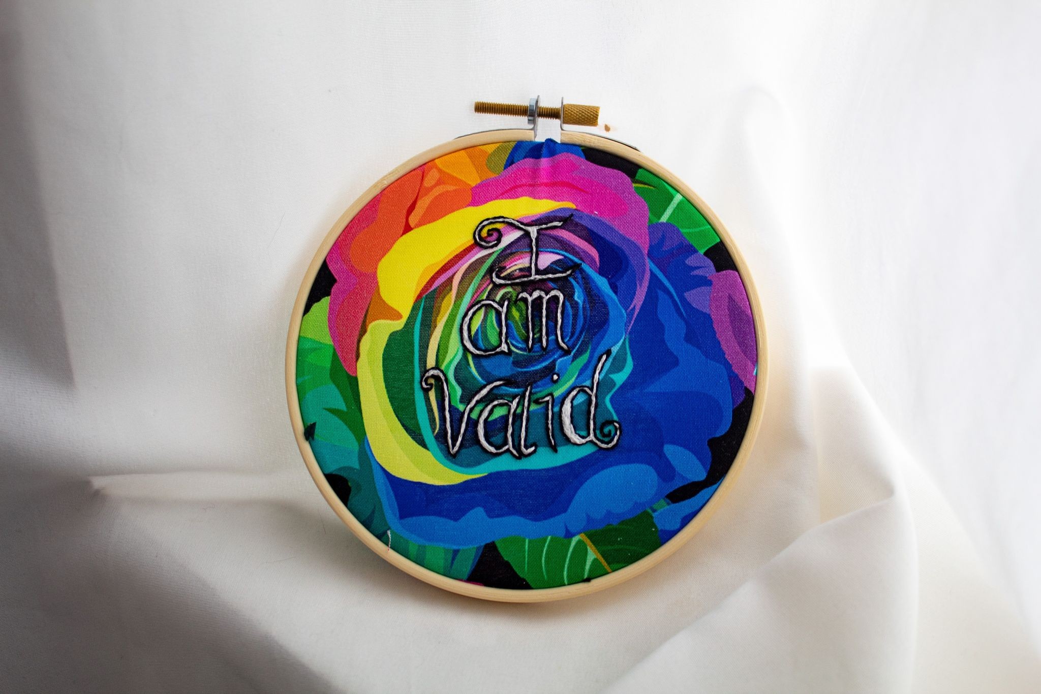 Product image of an embroidery featuring the words "I am valid"  in white thread with black outline embroidered over  rainbow-coloured rose-printed fabric, the whole in a wooden hoop and set against a white fabric backdrop