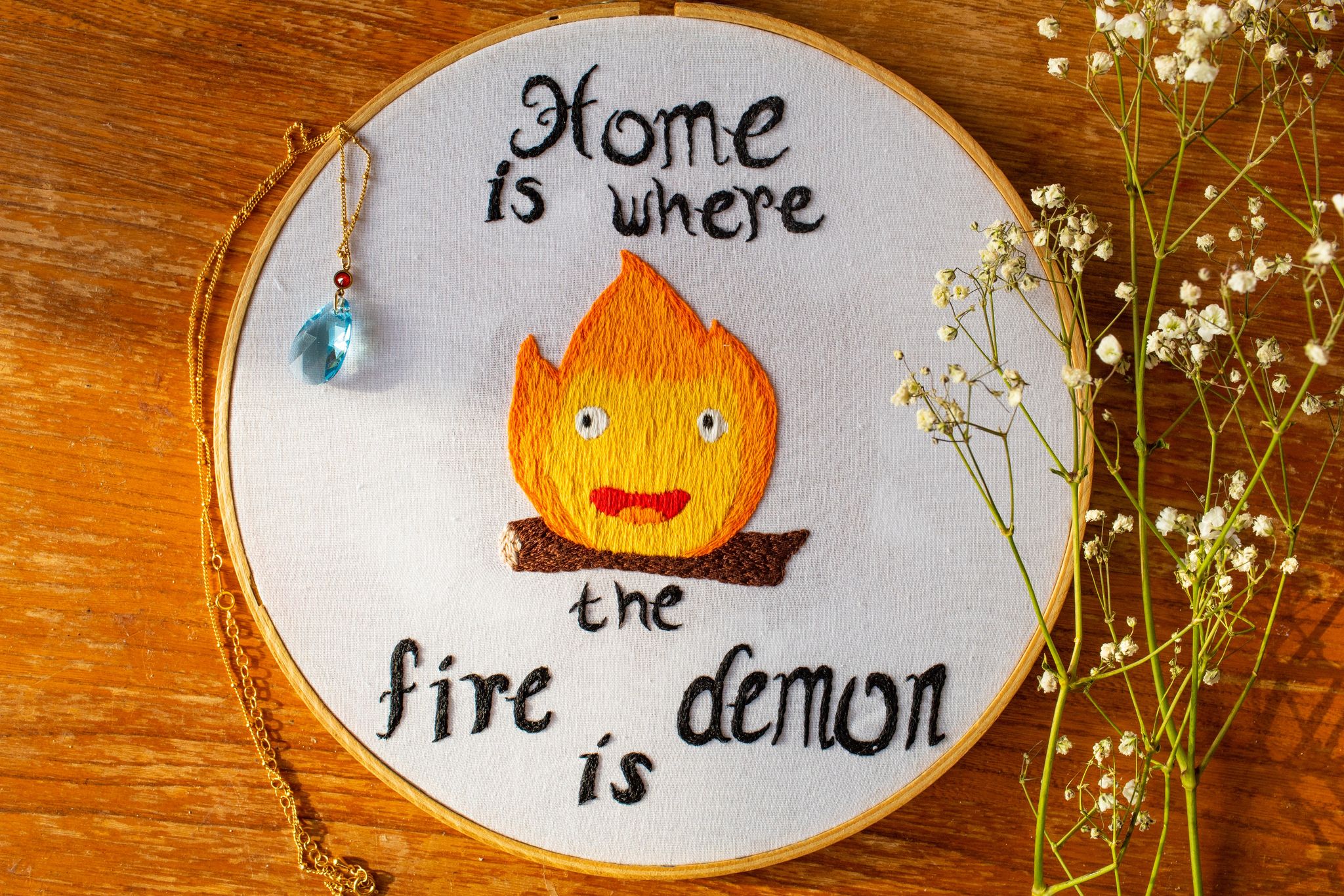 Product image of Calcifer embroider on wooden background surrounded by a blue pendant on a gold chain and baby's breath flowers