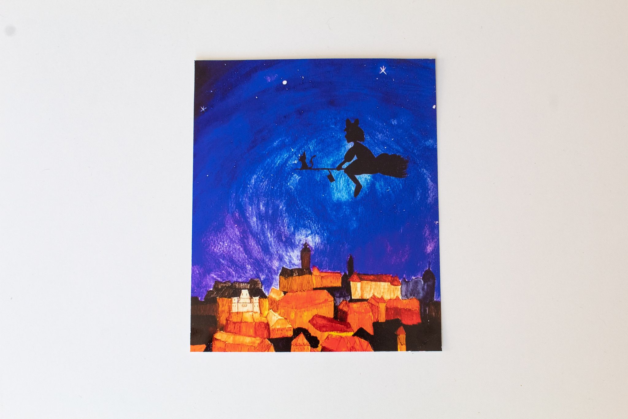 Product image of a print depicted a backlit witch girl atop a broom with a cat flying over an illuminated orange and yellow city, set against a dark blue and purple night sky