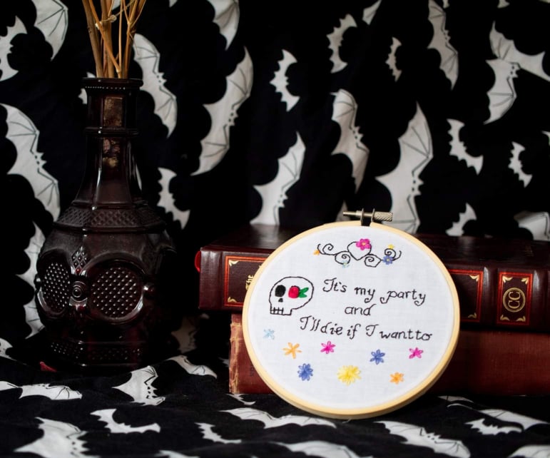 Product image of "It's My Party" embroidery set against black backdrop with white bats, leaning against old books and beside red gothic bottle holding dried red roses