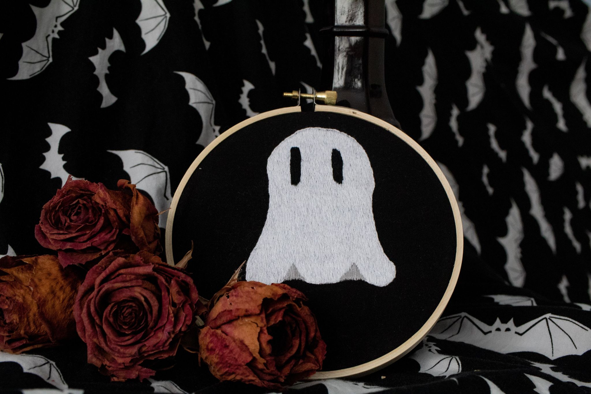 Product image of "Lonely Ghostie" embroidered ghost set against black backdrop with white bats, leaning on old books and surrounded by dried red roses