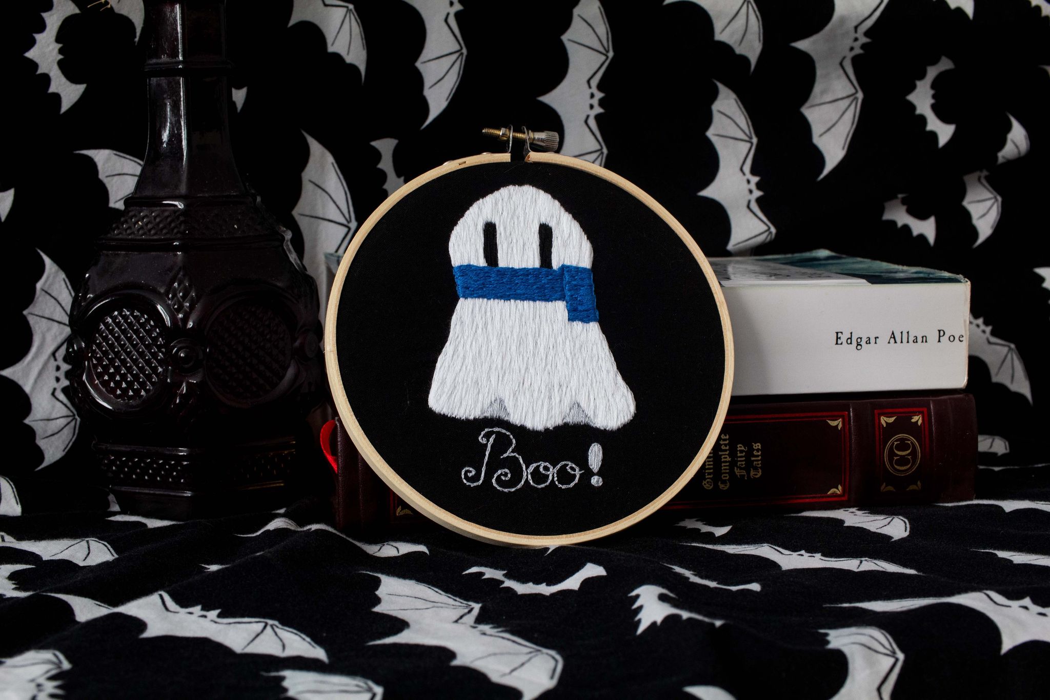 Product image of white embroidered ghost wearing a blue scarf on a black background with text "Boo!", set against a backdrop of old books and bats