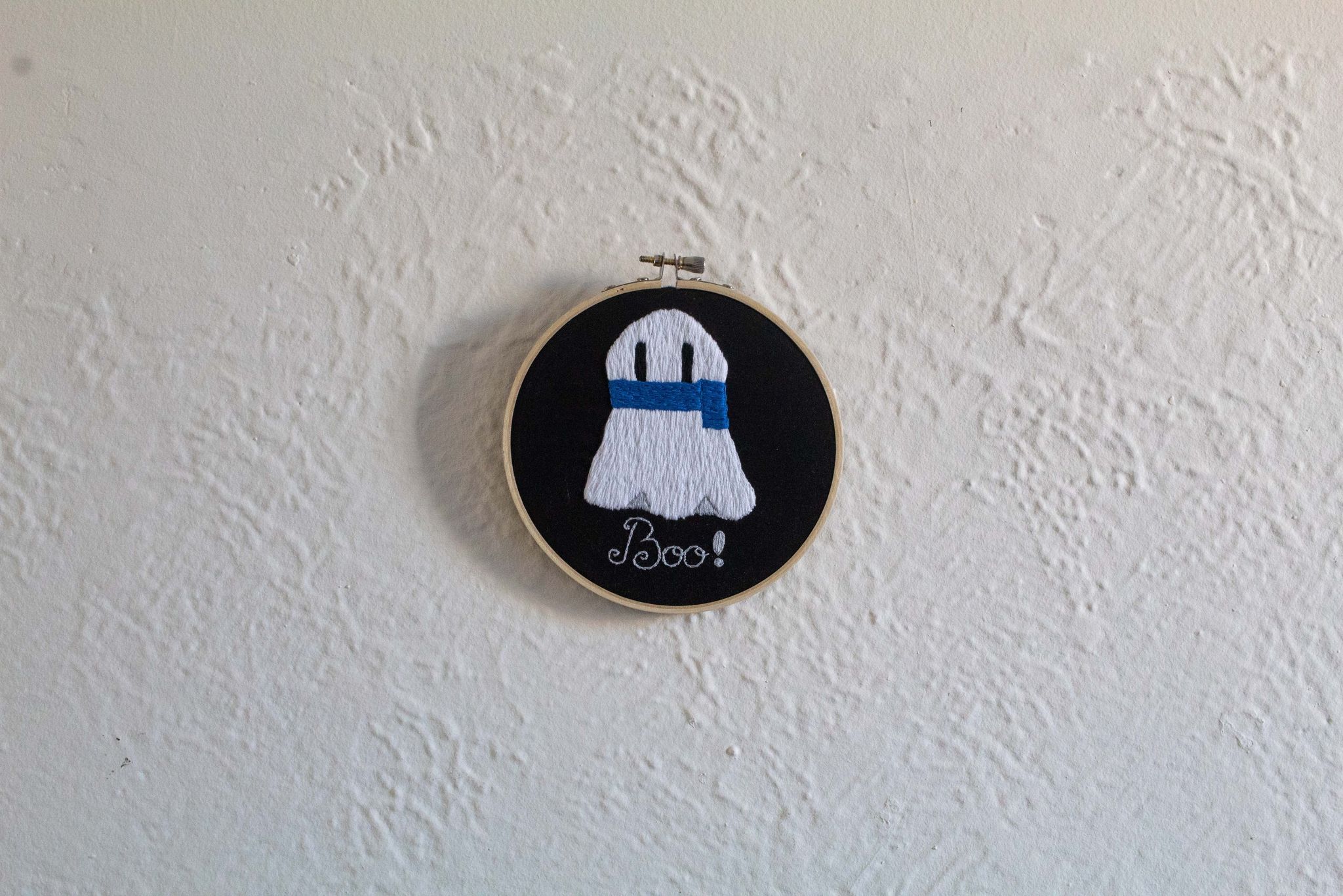 Product image of white embroidered ghost wearing a blue scarf on a black background with text "Boo!", hung up on a white wall