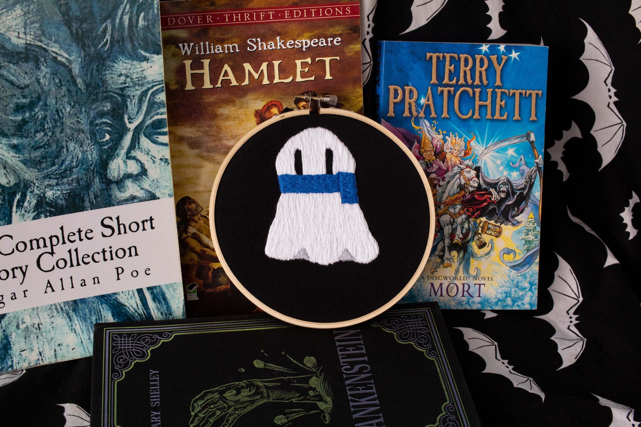 Product image of white embroidered ghost wearing a blue scarf on a black background, set against a backdrop of old books and bats
